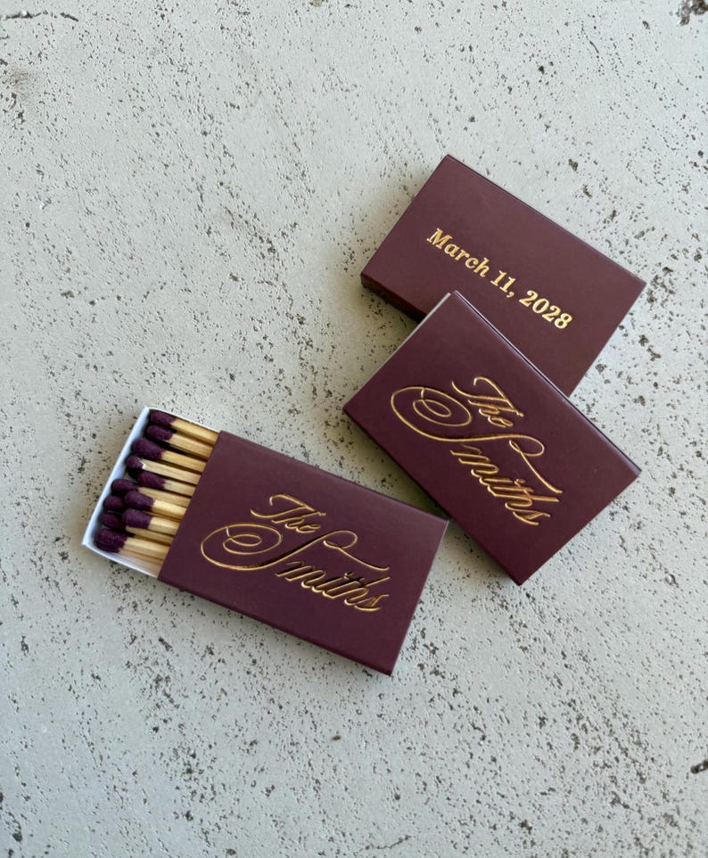May include: Three rectangular matchboxes in a deep burgundy color with gold foil lettering. Two boxes feature the script text The Smiths, while one box displays the date March 11, 2028. One box is partially open, revealing wooden matches with dark purple tips.
