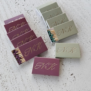 May include: A collection of rectangular matchboxes in deep plum and sage green, each featuring the gold embossed letters N&K in a cursive script. The plum boxes contain purple-tipped matches, while the sage boxes contain green-tipped matches.