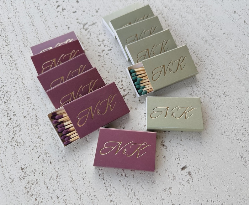 May include: A collection of rectangular matchboxes in deep plum and sage green, each featuring the gold embossed letters N&K in a cursive script. The plum boxes contain purple-tipped matches, while the sage boxes contain green-tipped matches.