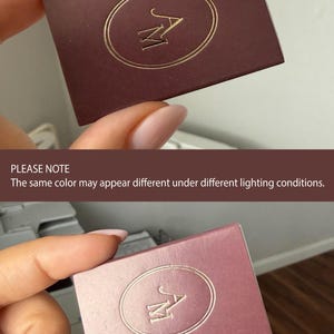May include: A rectangular box with a matte finish featuring a gold foil embossed oval frame and the letters AM in the center. The box is shown in two different shades, one a deep dark brown and the other a lighter mauve, to demonstrate color variation.