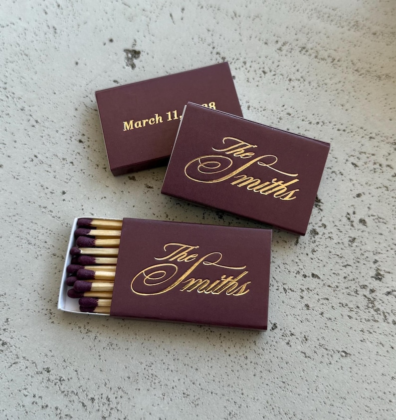 May include: Three rectangular matchboxes in deep burgundy with gold foil lettering. Two boxes feature the script text The Smiths, while one displays the date March 11, 1998. One box is partially open, revealing wooden matches with dark purple tips.