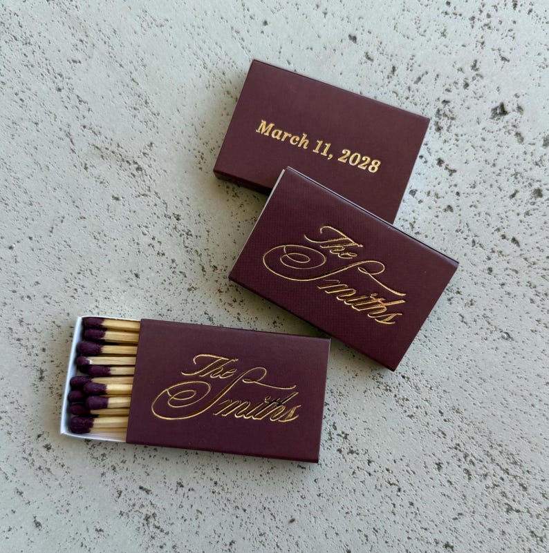May include: Three rectangular matchboxes in deep burgundy with gold foil lettering. Two boxes are closed, displaying the text "The Smiths" in script and "March 11, 2028" in print. One box is open, revealing wooden matches with dark purple tips.