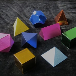 Puede incluir: Una colección de formas geométricas hechas de papel en varios colores, incluyendo rosa, azul, verde, dorado, plateado y rojo. Las formas incluyen un cubo, una pirámide, un dodecaedro y un prisma triangular.