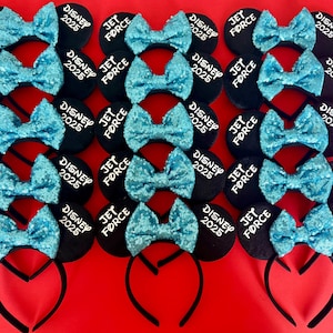 May include: A set of 12 black mouse ears with blue sequin bows. Each ear has white text that reads "JET FORCE" on one side and "DISNEY 2025" on the other side.
