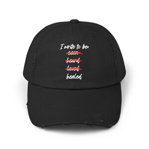 May include: Black distressed baseball cap featuring the text "I write to be: seen, heard, loved, healed" in white. The words "seen", "heard", and "loved" are crossed out in red. The cap has a curved peak and a worn appearance.