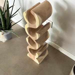 May include: A wooden wine rack with a unique, wavy design. The rack is made of light-colored wood and can hold multiple bottles. The design is modern and sculptural, perfect for displaying wine bottles. The rack is approximately 30 inches tall.