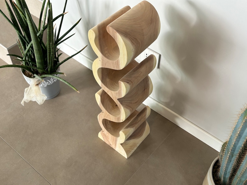 May include: A wooden wine rack with a unique, wavy design. The rack is made of light-colored wood and can hold multiple bottles. The design is modern and sculptural, perfect for displaying wine bottles. The rack is approximately 30 inches tall.