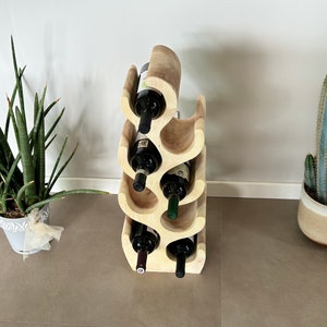 May include: A wooden wine rack with a unique wave design, holding six bottles of wine. The rack is light brown and has a natural wood finish. The rack is standing on a light brown tiled floor.