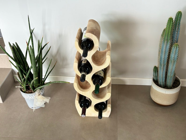May include: A wooden wine rack with a unique wave design, holding six bottles of wine. The rack is light brown and has a natural wood finish. The rack is standing on a light brown tiled floor.