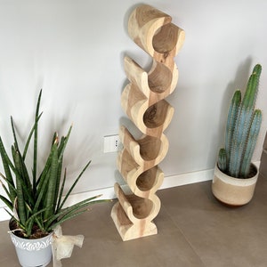 May include: A wooden wine rack with a unique, curved design. The rack is made of light-colored wood and features six individual slots for wine bottles. The rack is free-standing and has a natural, organic aesthetic, perfect for home decor.