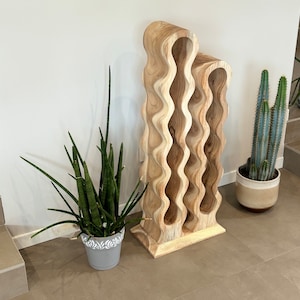 May include: A wooden wine rack with a wavy design. The rack is made of light brown wood and has six compartments. The rack is standing on a light brown tile floor.