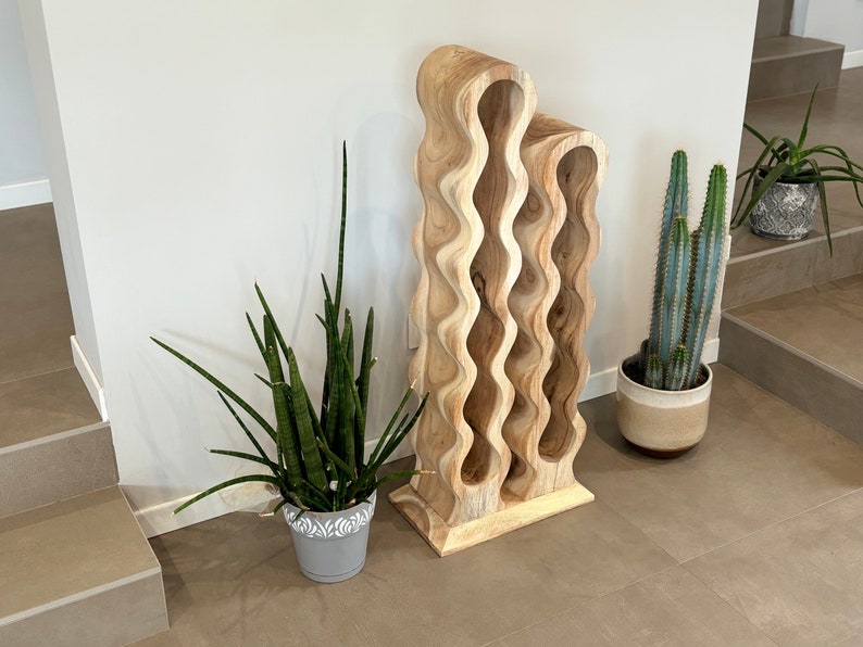 May include: A wooden wine rack with a wavy design. The rack is made of light brown wood and has six compartments. The rack is standing on a light brown tile floor.