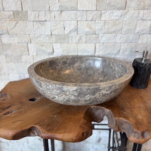 Peut inclure: Un lavabo en pierre grise de forme arrondie. Le lavabo est posé sur une table en bois naturel avec un bord brut. Un distributeur de savon noir est visible à droite. L'arrière-plan présente un mur de briques de couleur claire.