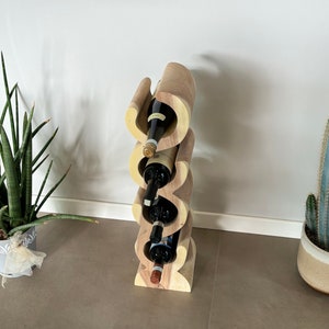 May include: A wooden wine rack with a unique, wavy design, holding four wine bottles. The rack is made of light and dark wood tones. The image also shows potted plants, including a snake plant and a cactus, adding a touch of greenery to the scene.