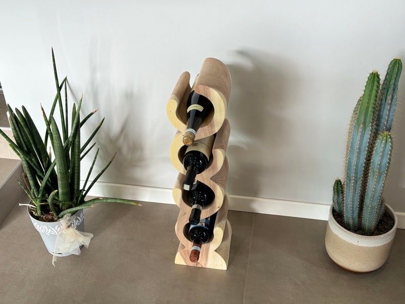 May include: A wooden wine rack with a unique, wavy design, holding four wine bottles. The rack is made of light and dark wood tones. The image also shows potted plants, including a snake plant and a cactus, adding a touch of greenery to the scene.