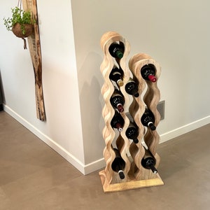May include: A wooden wine rack with a wavy design holds multiple bottles of wine. The rack is made of light brown wood and has a natural finish. The bottles are arranged in a staggered pattern, with the tops of the bottles visible.