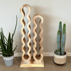 May include: A wooden wine rack with a wavy design. The rack is made of two pieces, one taller than the other. The rack is standing on a light brown floor. There are two potted plants in the background, one with green leaves and the other with a cactus.