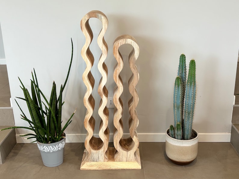 May include: A wooden wine rack with a wavy design. The rack is made of two pieces, one taller than the other. The rack is standing on a light brown floor. There are two potted plants in the background, one with green leaves and the other with a cactus.