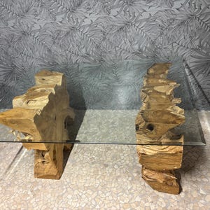 May include: A contemporary coffee table featuring a clear glass surface resting on two bases crafted from natural wood. The bases, made of light brown, textured wood, provide a unique, organic aesthetic. The table is a statement piece.