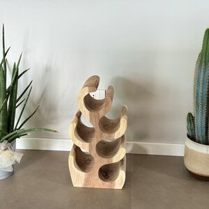 May include: A wooden wine rack shaped like a tree with six bottle holders. The rack is made of light brown wood and has a natural finish. The rack is sitting on a light brown floor.