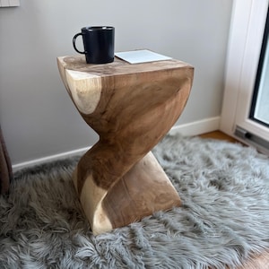 Puede incluir: Una mesa auxiliar de madera natural con un diseño retorcido único. La mesa tiene una taza negra y un pequeño cuadrado blanco encima. La mesa está sobre una alfombra gris.