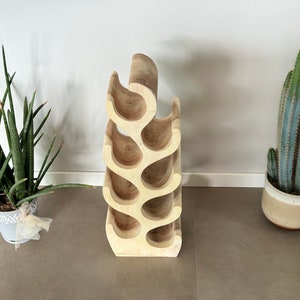 May include: A wooden wine rack shaped like a tree branch with six bottle holders. The rack is light brown and has a natural wood finish. It is sitting on a light brown tiled floor.