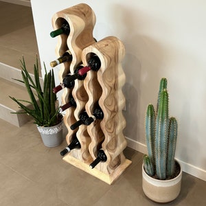 May include: A wooden wine rack with a wavy design, holding multiple bottles of wine. The rack is made of light brown wood and has a natural finish. The rack is standing on a light brown tiled floor.