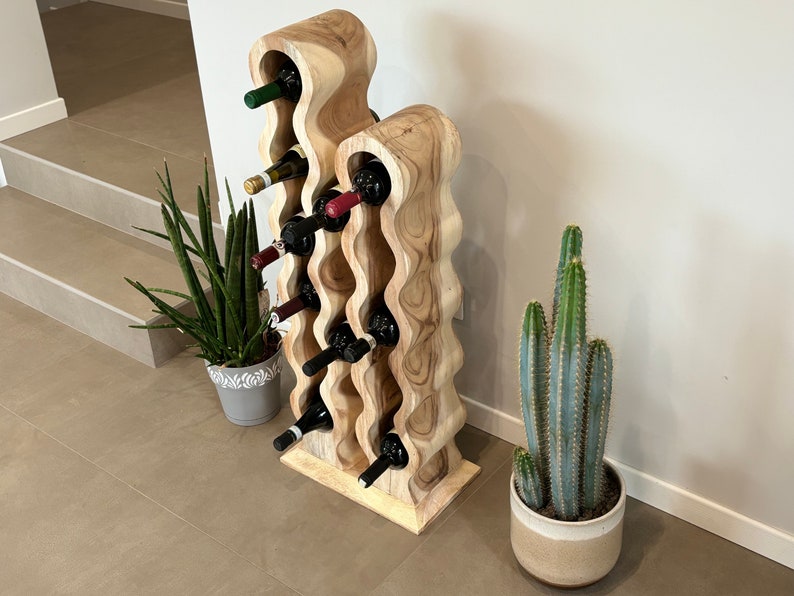 May include: A wooden wine rack with a wavy design, holding multiple bottles of wine. The rack is made of light brown wood and has a natural finish. The rack is standing on a light brown tiled floor.