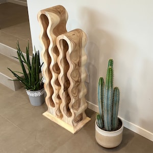 May include: A wooden wine rack with a unique wavy design. The rack is made of light brown wood and has 10 slots for bottles. The rack is standing on a light brown tiled floor.