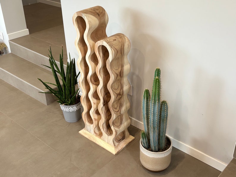 May include: A wooden wine rack with a unique wavy design. The rack is made of light brown wood and has 10 slots for bottles. The rack is standing on a light brown tiled floor.