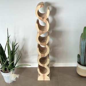 May include: A tall, wooden wine rack with a natural finish. The rack has a unique, curved design with multiple openings to hold wine bottles. The rack is surrounded by potted plants, including a snake plant and a cactus.