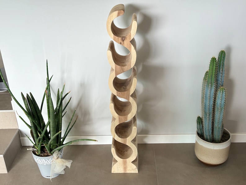 May include: A tall, wooden wine rack with a natural finish. The rack has a unique, curved design with multiple openings to hold wine bottles. The rack is surrounded by potted plants, including a snake plant and a cactus.