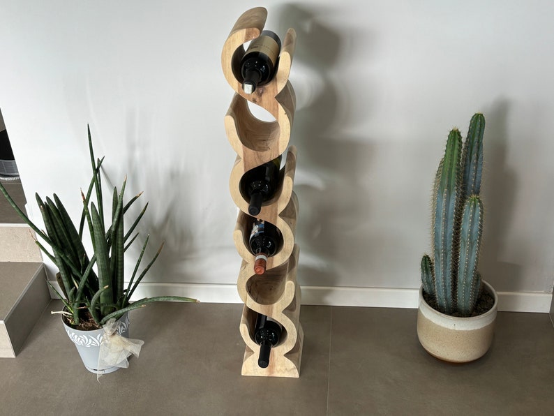May include: A wooden wine rack with a unique, curved design, holding several wine bottles. The rack is made of light-colored wood and stands vertically. The image also shows potted plants, including a snake plant and a cactus.