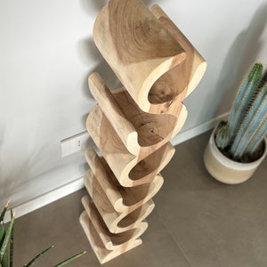 May include: A wooden wine rack with a unique, wavy design. The rack is made of light-colored wood and has six openings to hold wine bottles. The rack is positioned against a white wall, with a cactus in a pot visible in the background.