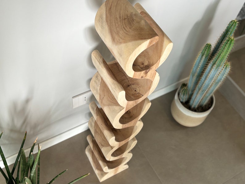 May include: A wooden wine rack with a unique, wavy design. The rack is made of light-colored wood and has six openings to hold wine bottles. The rack is positioned against a white wall, with a cactus in a pot visible in the background.