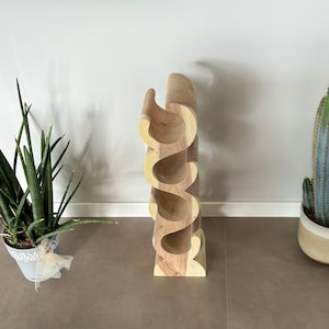 May include: A wooden wine rack with a unique, curved design. The rack is made of light-colored wood and can hold several bottles. A potted cactus and a snake plant are on either side of the wine rack.