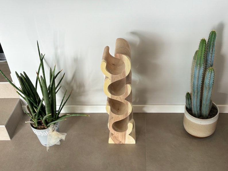 May include: A wooden wine rack with a unique, curved design. The rack is made of light-colored wood and can hold several bottles. A potted cactus and a snake plant are on either side of the wine rack.