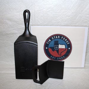 May include: Black cast iron spatula and lid lifter with a handle and a logo that says "Tin Star Forge EST 2022" in a red, white, and blue circle. The spatula has a rectangular blade and a loop handle.