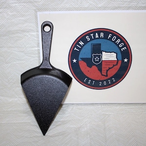 May include: A black cast iron pizza peel with a triangular blade and a handle with a hanging hole. The peel is next to a circular logo with the text "TIN STAR FORGE" and "EST 2022" with a Texas outline.