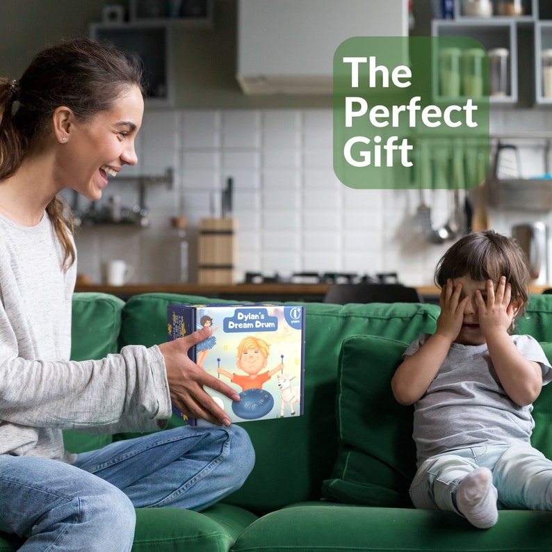 May include: A woman smiles while presenting a personalized drum set to a child who is covering their eyes with their hands. The box features a colorful illustration of a child playing the drum with the text "Dylan's Dream Drum". The text "The Perfect Gift" is displayed in a green circle.