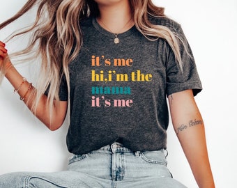 May include: Dark gray t-shirt with the text "it's me hi, i'm the mama it's me" in colorful lettering. The shirt is paired with light wash jeans and white sneakers. The overall style is casual and comfortable.
