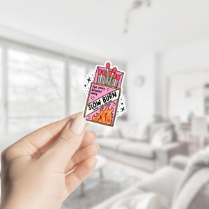 May include: A sticker featuring a pink matchbox with the words "SLOW BURN book club matches" and flames. The matchbox also has the text "for spicy bitches only". The matches are red. The sticker is held by a hand.