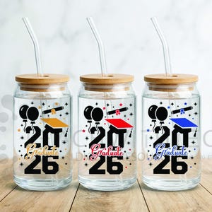 Op de afbeelding: Drie heldere glazen bekers met bamboe deksels en rietjes. Elke beker heeft een afstudeerthema-ontwerp met het jaartal "26" en het woord "Graduate". De ontwerpen omvatten ballonnen, afstudeerpetten en diploma's in verschillende kleuren.