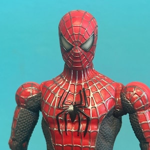 May include: A red and black action figure of a superhero character with a full-body mask, large white eyes, and a prominent black spider emblem on the chest. The suit features a detailed silver web pattern across the red sections and textured black panels on the sides and arms.