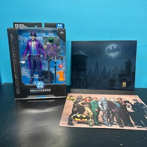 May include: A McFarlane Collector Edition Multiverse action figure of The Joker in a purple coat and plaid pants, packaged in a box with accessories. Beside it is a backdrop card showing a city skyline with a bat signal and a character art print.