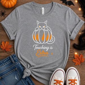 May include: A heather gray short-sleeve t-shirt featuring a white line-art graphic of a cat sitting behind an orange pumpkin. Below the graphic, the text reads "Touching is Extra" in white and orange script and sans-serif lettering.