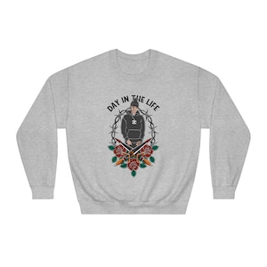 May include: Gray sweatshirt with a graphic of a person wearing a black hoodie and a black cap with a white logo. The graphic is surrounded by a barbed wire circle with the text "DAY IN THE LIFE" above it and two crossed knives with red roses below it.