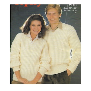 Könnte beinhalten: Zwei Personen tragen weiße Strickpullover mit V-Ausschnitt. Die Pullover haben eine gerippte Struktur. Der Text "Aran" und "Größen 32"-44" 81cm-112cm" ist im Bild sichtbar.