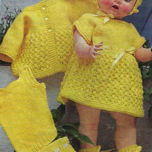 Könnte beinhalten: Eine Vintage-Puppe in einem handgefertigten, gelb gestrickten Outfit, einschließlich Kleid, Kapuze, Strickjacke, Weste und Stiefeletten. Die Puppe hat rosige Wangen und posiert mit den Kleidungsstücken. Die Strickartikel haben ein strukturiertes Muster.