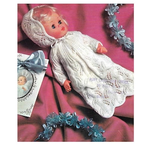 Puede incluir: Un vestido y un gorro de bebé para muñeca de crochet blanco con un patrón de encaje. La muñeca está tumbada sobre una superficie rosa con adornos de flores azules.